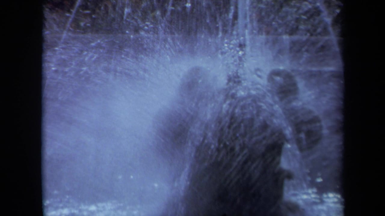 1969 California 8mm frame showing a close, blue-tinted view of fountain spray with distant park visitors and trees behind it.