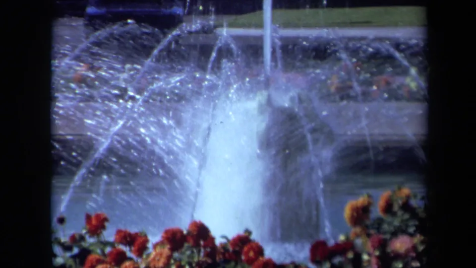 1969 California 8mm frame showing a wider park fountain view with arcing water, blooming flowers, grass, and background cars.