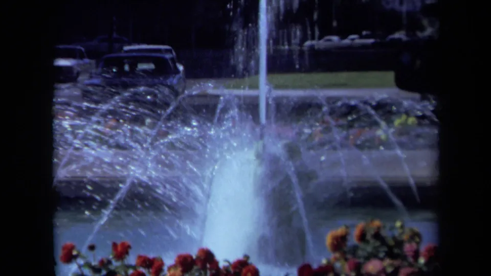 Late-1960s California home-movie frame showing fountain water arcing over flowers, with grass and parked cars in the background.