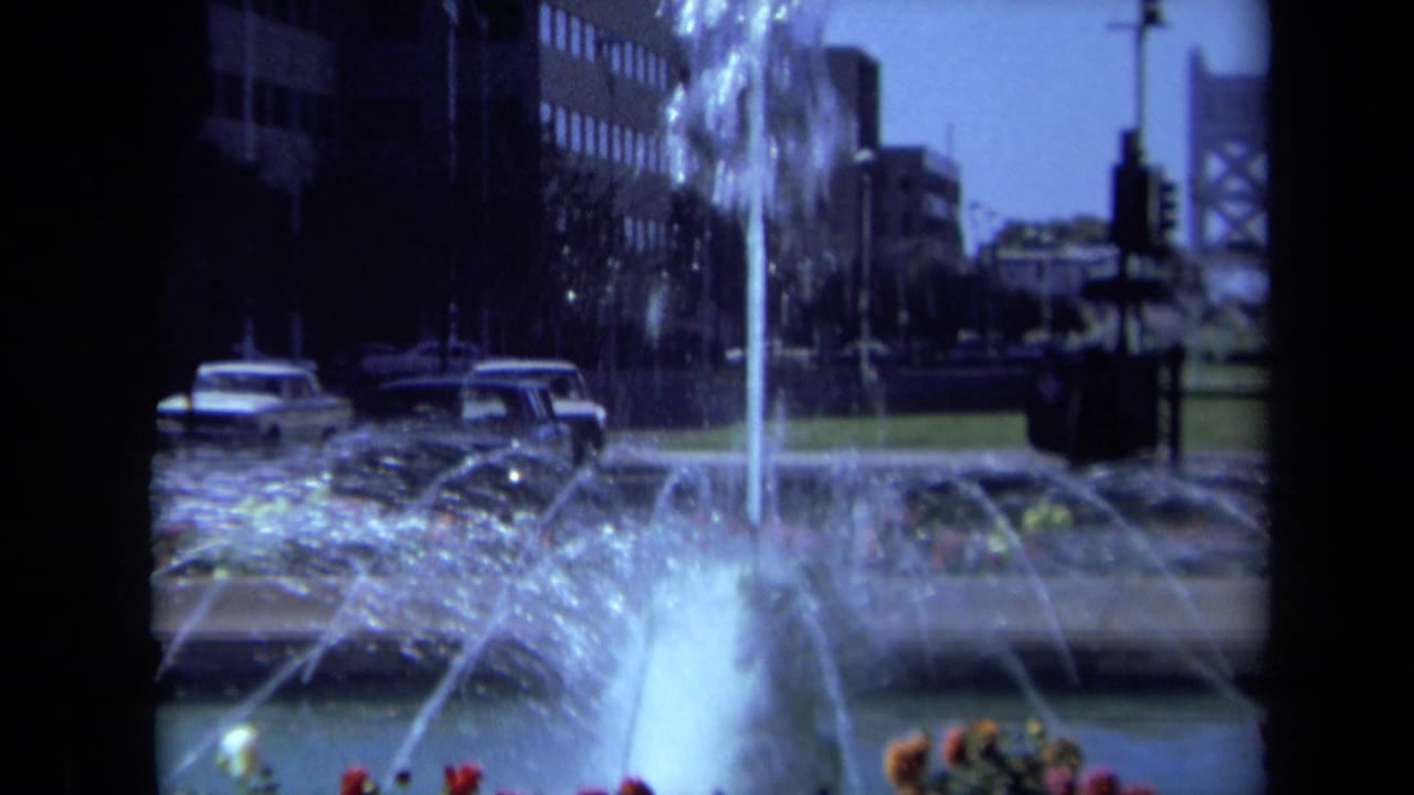 1969 California 8mm frame showing a city fountain with water jets, flower beds, parked cars, roads, trees, office buildings, and a distant bridge-like form.