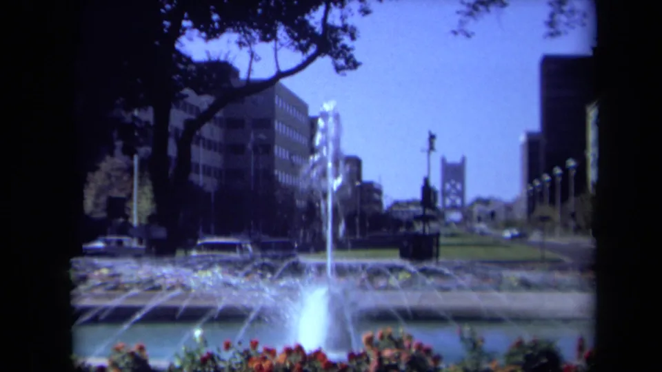 Wider 1969 California home-movie view of a landscaped urban fountain with trees overhead, office buildings, cars, lawn, and a distant bridge-like structure.