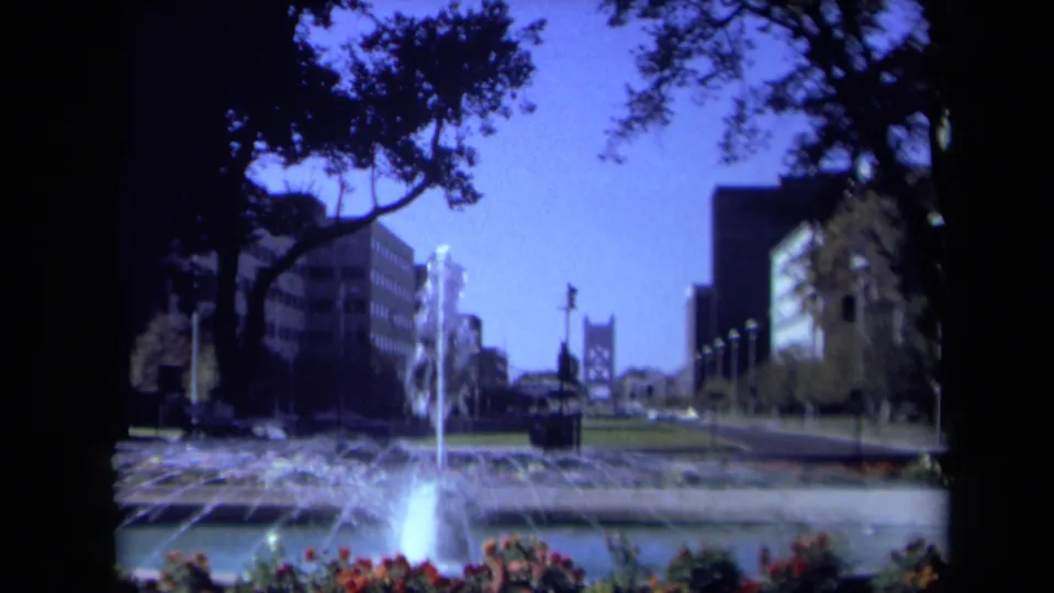 1969 California 8mm frame with the fountain lower in view, flower beds at the edge, trees above, office buildings, roads, and a distant bridge-like form.