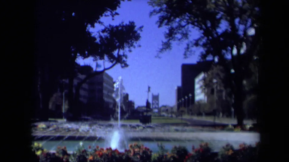 Wide 8mm view of a California city fountain in 1969, with landscaped planting, shade trees, and modern buildings across the background.