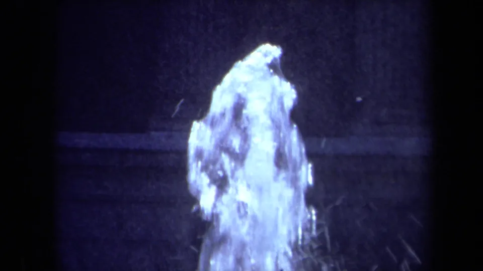 Close 1969 California home-movie view of fountain water rising and splashing, with the pool edge and dark shadowed surface behind it.