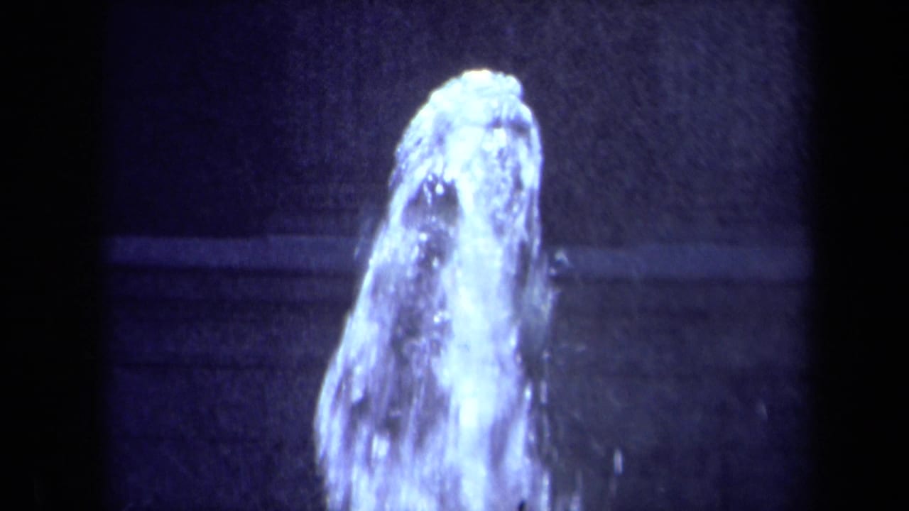 Close 1969 8mm view of a fountain jet or water spray in a California urban plaza, framed by dark architectural shadow.