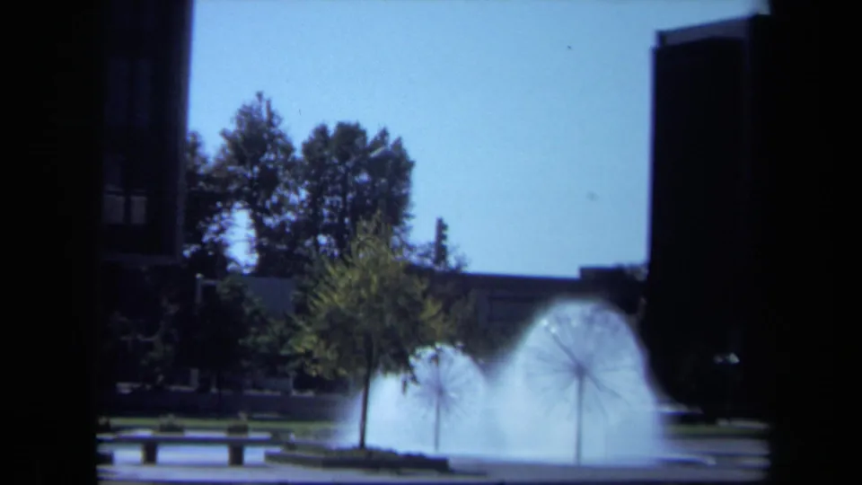 1969 California home-movie view of a public fountain and plaza, with trees and benches framed by a dark vertical structure.