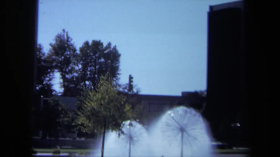 Late-1960s California plaza view showing a dandelion-shaped fountain beside a small tree, with dark buildings at the edges.