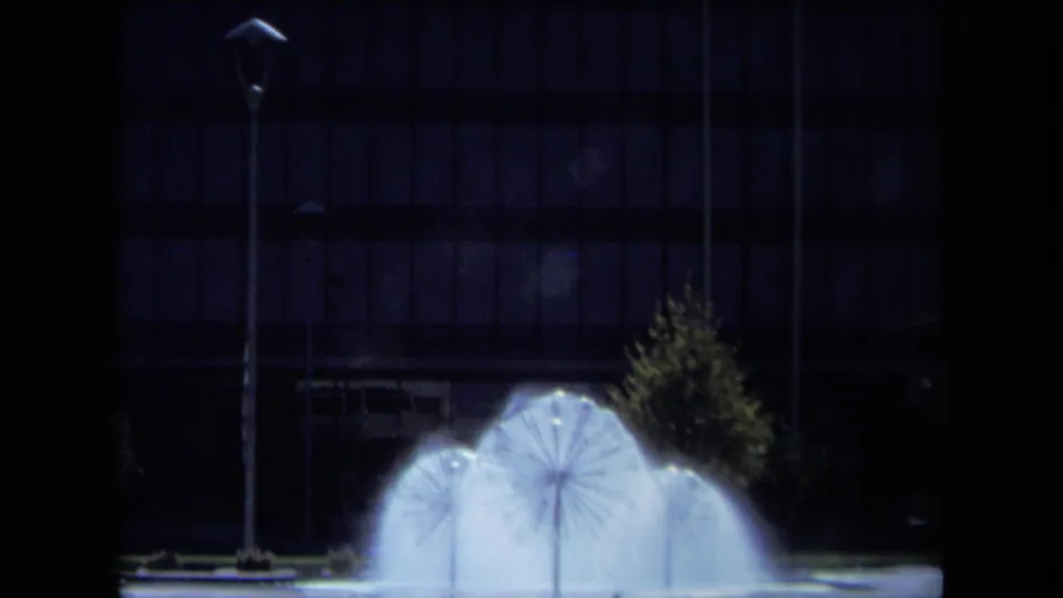 Daylight 8mm view of the fountain spraying beside a small tree, street lamp, and dark grid-window building in California.