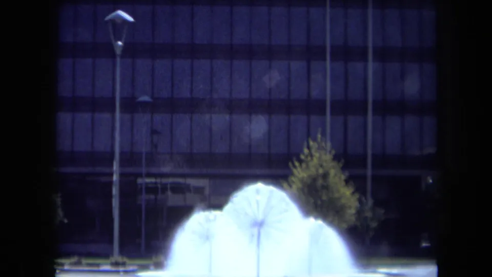 Nighttime 1969 California frame showing the illuminated fountain below slim street lamps and a modern building facade.