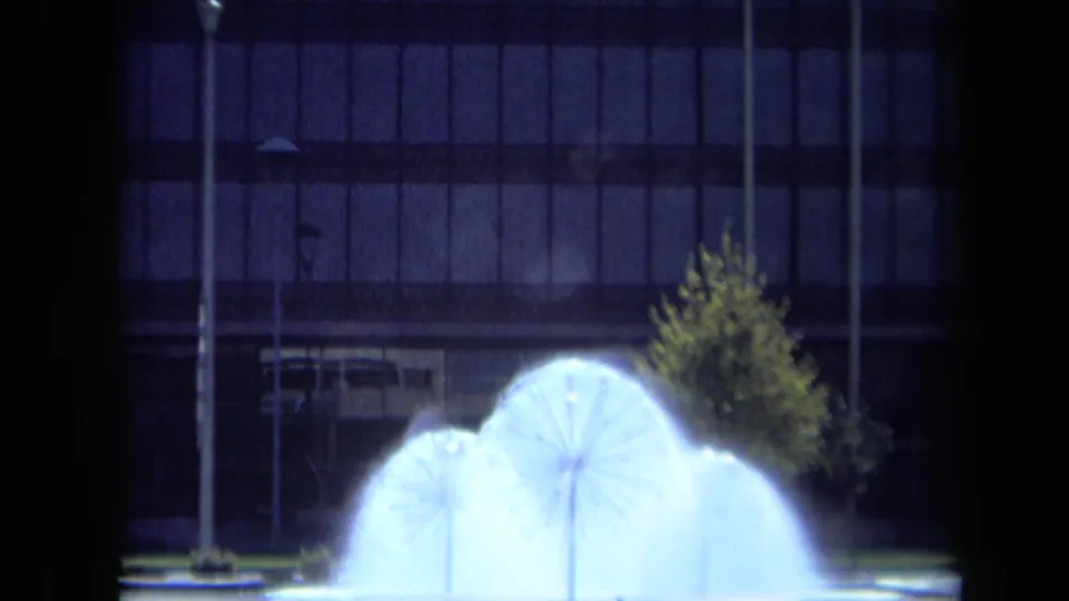 Night 8mm frame of the dandelion-shaped fountain glowing in front of a small tree, street lamp, and grid-window building.