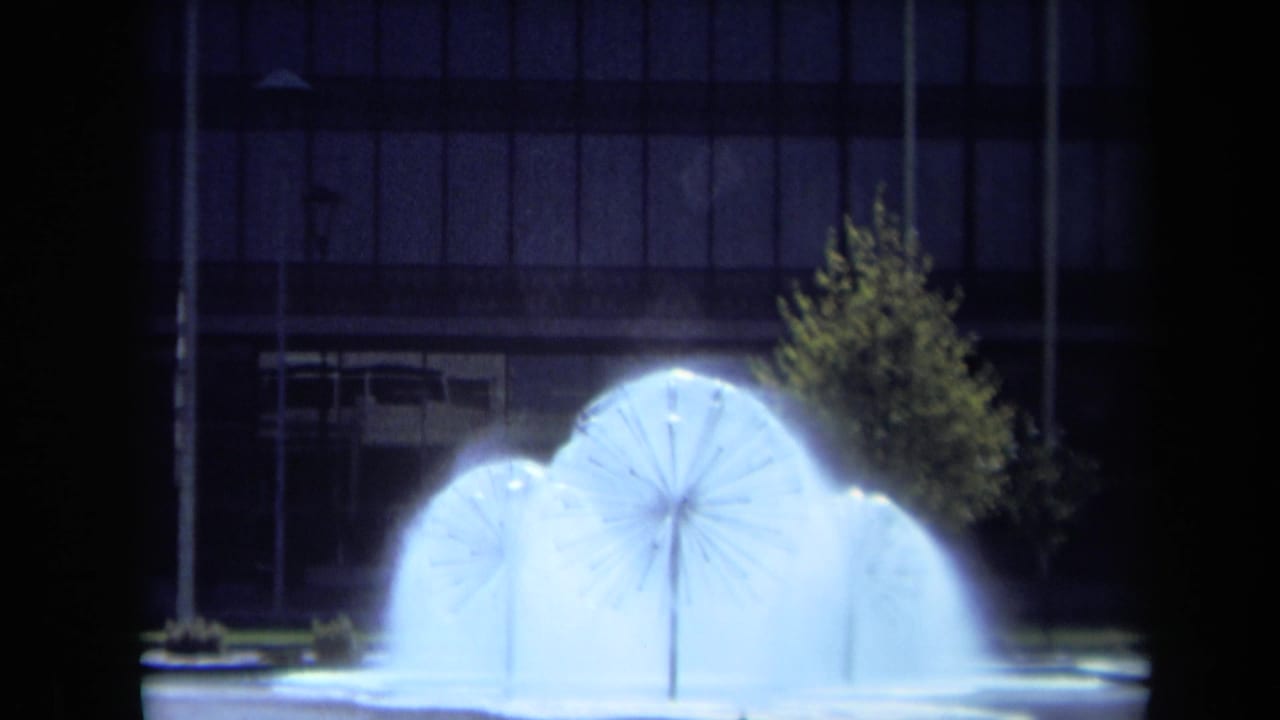Three illuminated water fountains spray in circular patterns before a dark building and tree.