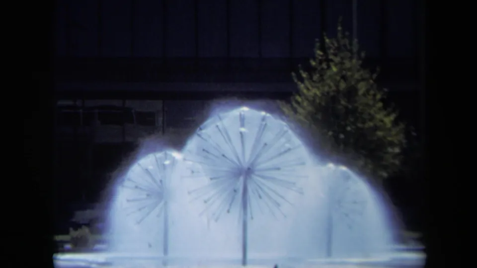 Nighttime 8mm view of three glowing circular fountains in California, framed by a dark building facade and a tree.