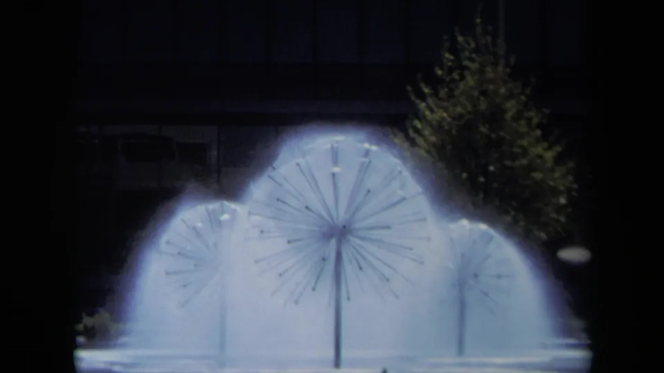 Close 1969 California frame of blue-white fountain spray at night, with a tree and dark modern building behind it.