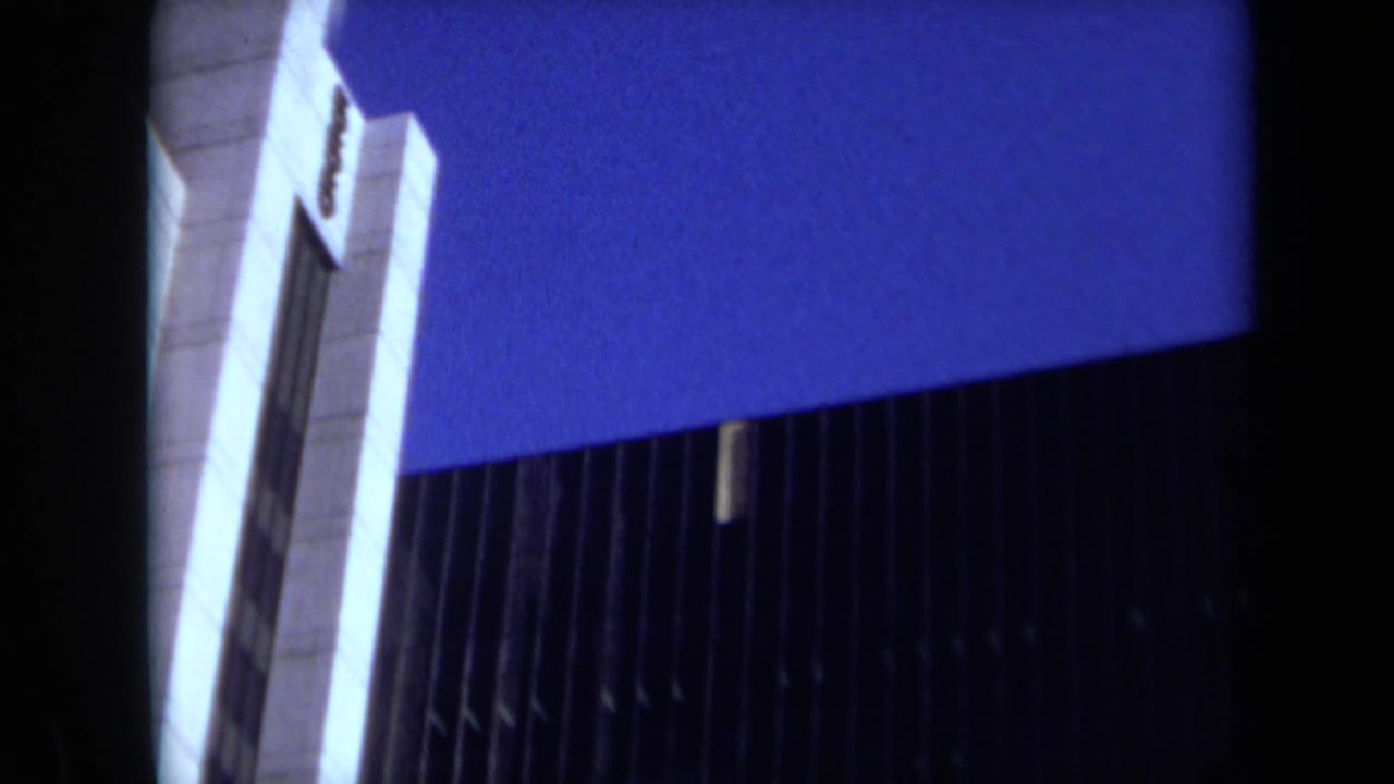 1969 California home-movie frame showing a low-angle view of a modern skyscraper with vertical facade ribs and blue twilight sky.