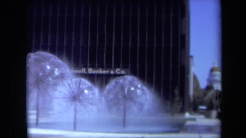1969 California 8mm frame of multiple kinetic fountain forms spraying water before a dark commercial building.