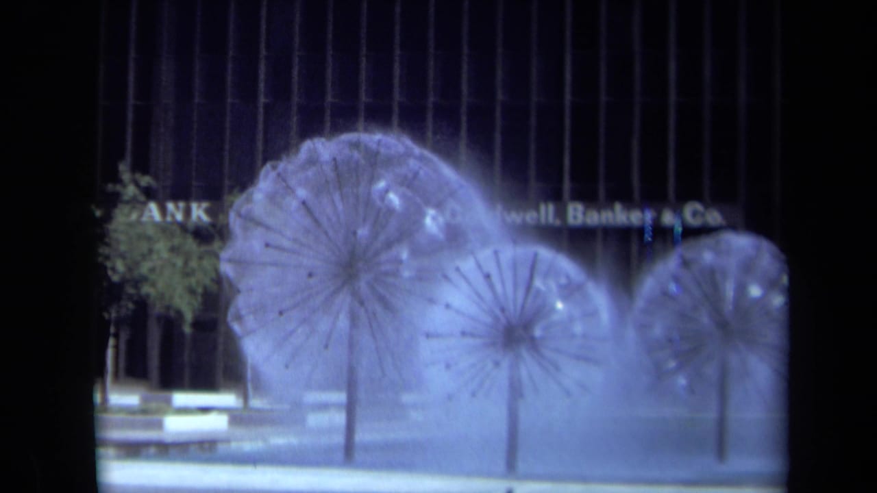 1969 California 8mm frame showing three sculptural water fountains before a modern facade with partial Coldwell Banker & Co. signage.