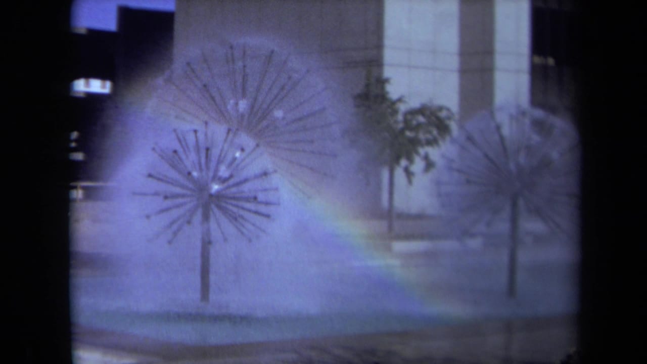 1969 Sacramento 8mm frame showing spiky metal fountains spraying mist in a public plaza as a rainbow appears across the water.
