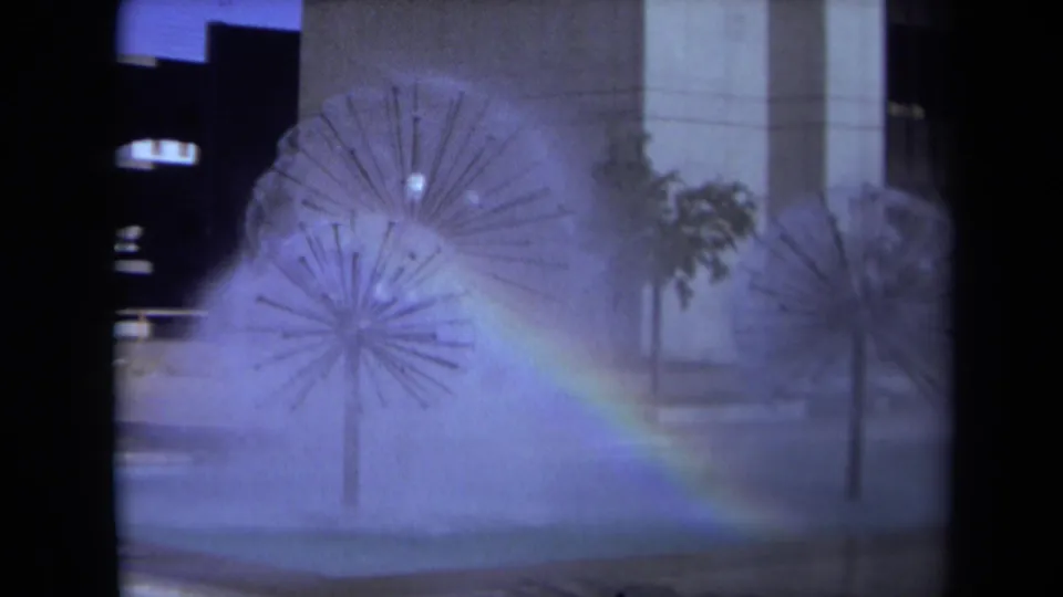 1969 Sacramento home-movie frame with dandelion-like fountains, drifting spray, a rainbow, a tree, and modern building facades.