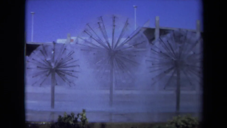 1969 Sacramento home-movie frame of three spiky public fountains spraying mist against a blue evening sky and modern plaza buildings.