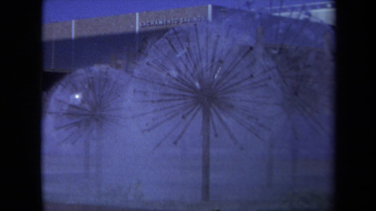 1969 8mm Sacramento view of rain falling on spherical dandelion-like sculptures near a Sacramento Savings sign.