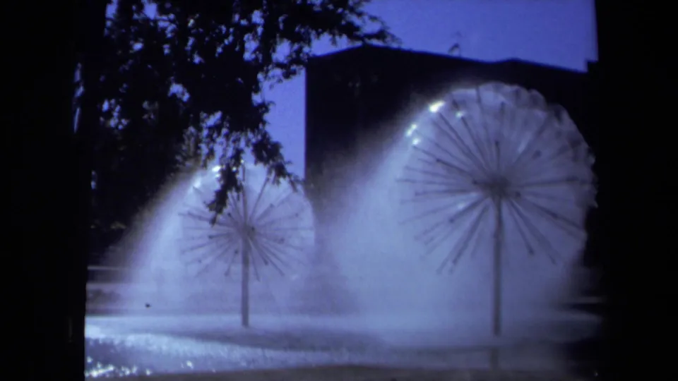 1969 Sacramento 8mm frame of two starburst fountain sprays and bright mist, partly framed by tree canopy and a dark background structure.