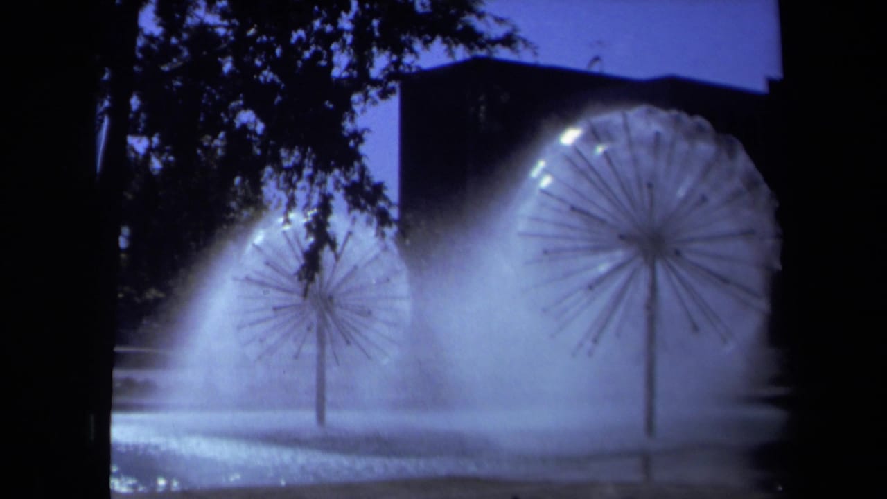 1969 Sacramento 8mm frame showing glowing dandelion-like sprinklers spraying misty arcs at night near trees and a building.