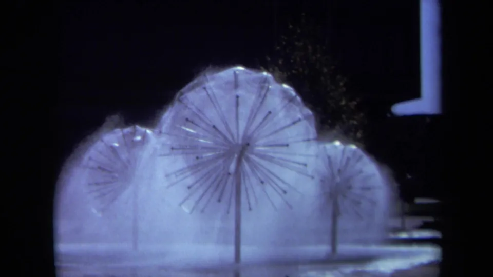 1969 Sacramento frame showing a prominent illuminated sprinkler sculpture with mist, wet ground, and dark architectural background.