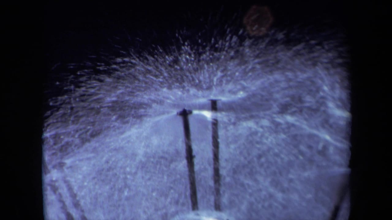 Close-up of black sprinkler poles spraying water across a pale blue-tinted surface.