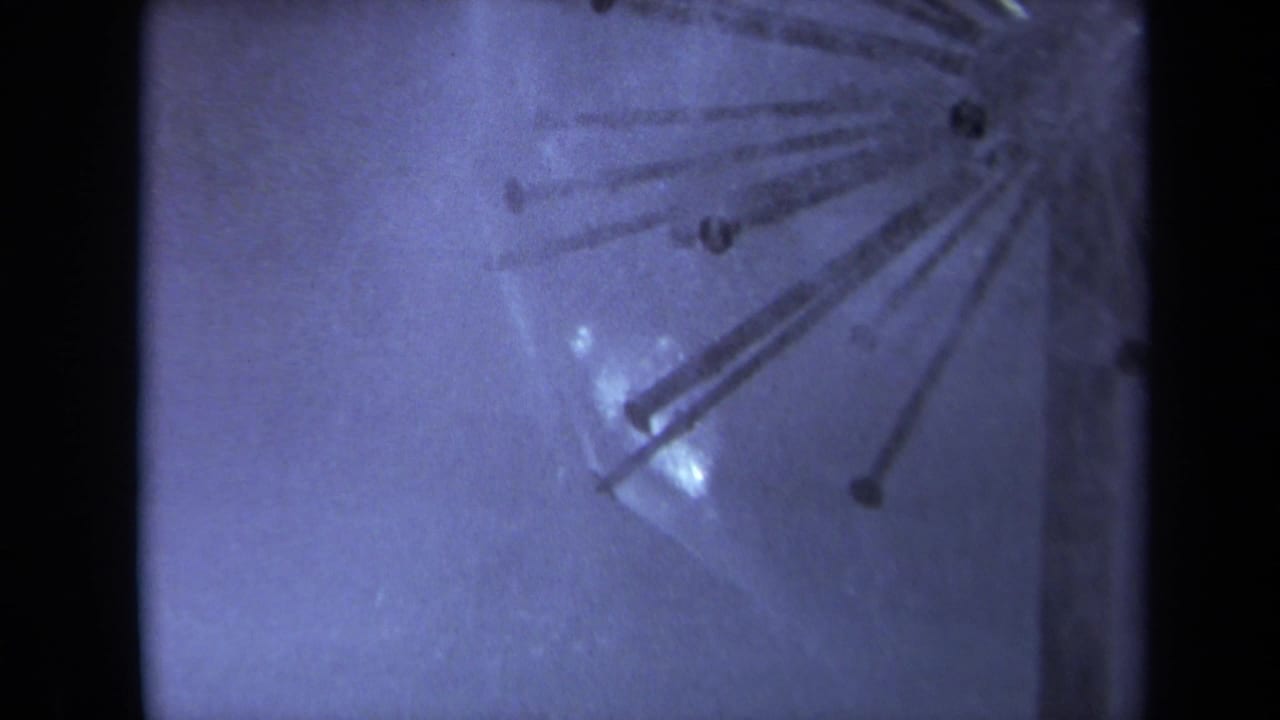 Blue-tinted close-up of wet surface with radial metal rods and dark film edges.