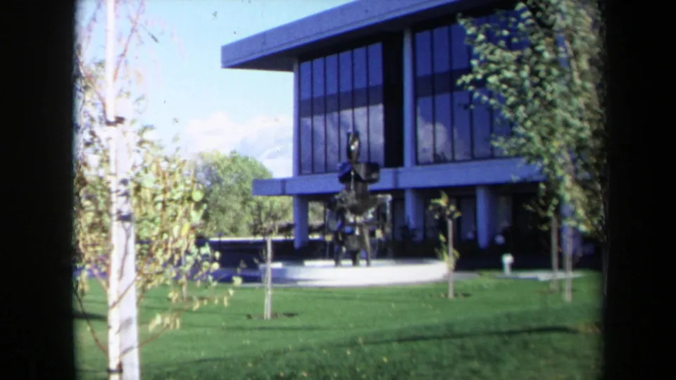1969 Sacramento 8mm view of a modernist glass-fronted building with lawn, young trees, and an abstract sculpture.