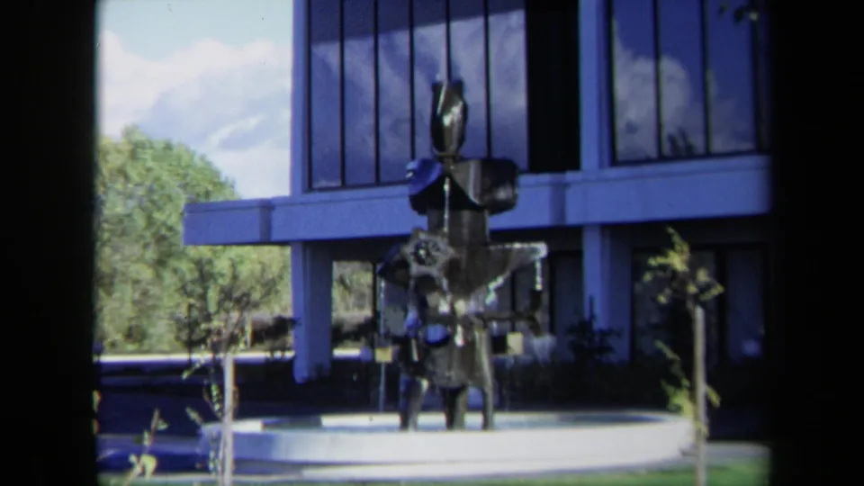 Closer 1969 Sacramento home-movie view of the dark metal sculpture, pale circular base, and modern glass office exterior.