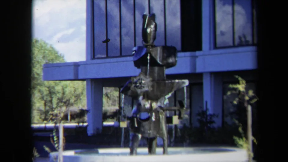 Tight 1969 Sacramento frame of the abstract metal fountain sculpture with reflective blue office windows and trees behind it.
