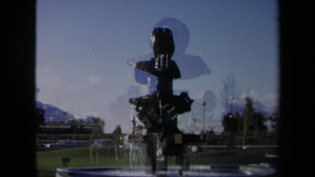 1969 Sacramento 8mm frame showing a silhouetted modernist fountain, water spray, grass, trees, and distant buildings.