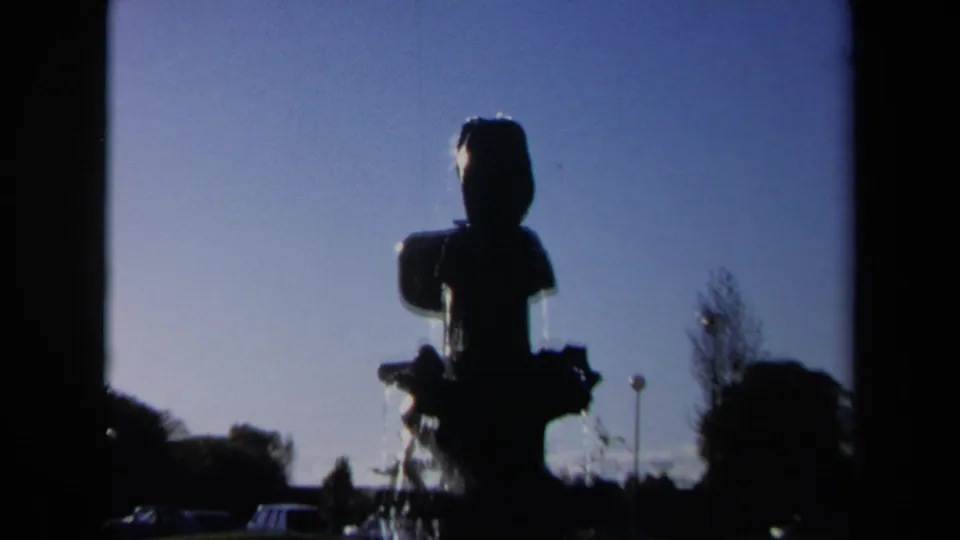 1969 Sacramento frame of the dark geometric fountain seen from farther back, with blue sky, trees, lamp posts, and water spray.