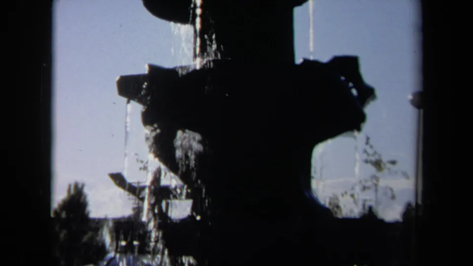 The 8mm frame shows the fountain in silhouette above a green lawn, with water falling and trees visible beyond the public outdoor setting.
