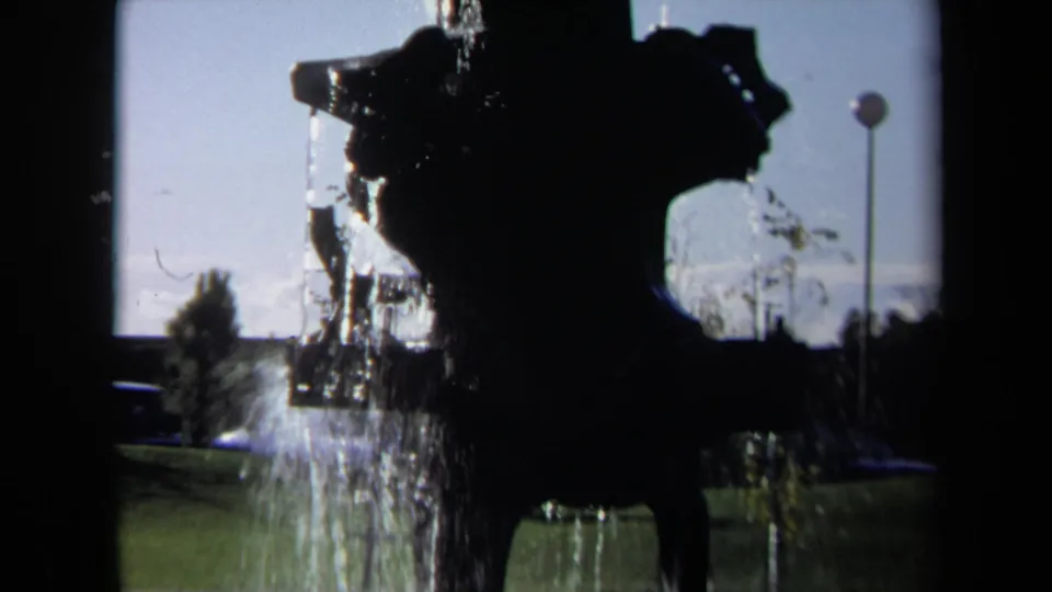 A 1969 Sacramento home-movie frame shows the fountain's dark profile, falling water, green lawn, distant trees, and a round-topped pole at right.