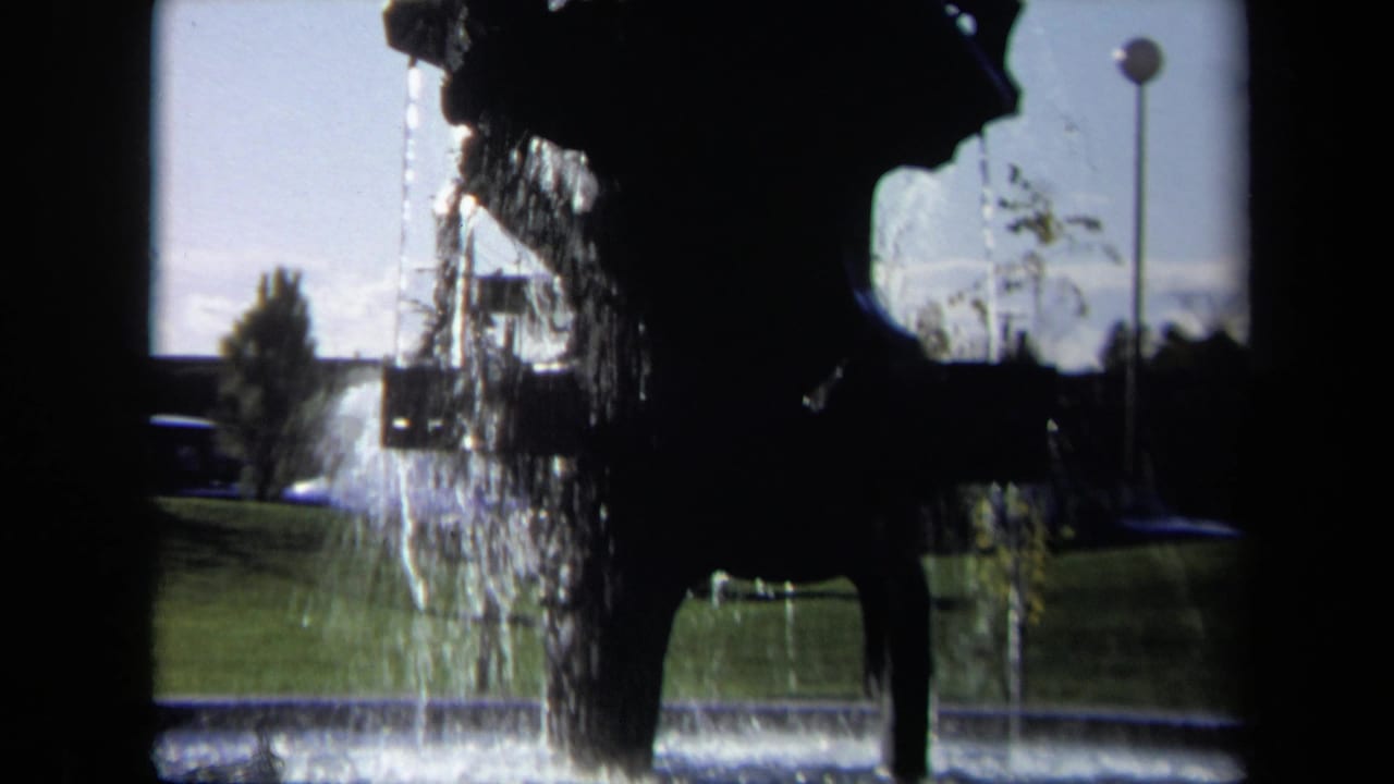 1969 Sacramento home-movie frame of a silhouetted fountain sculpture with cascading water, grass, road edge, and pole light in the background.