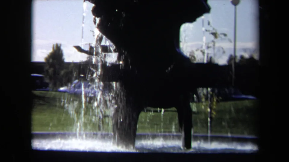1969 Sacramento frame showing the fountain sculpture in strong silhouette as water spills across the basin, with grass and distant cars beyond.