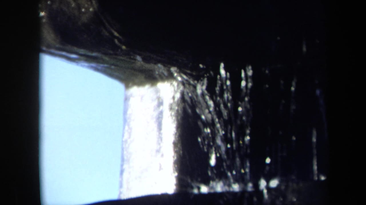 Close 8mm view of water cascading over dark metal mill parts, with a pale blue opening behind the structure.
