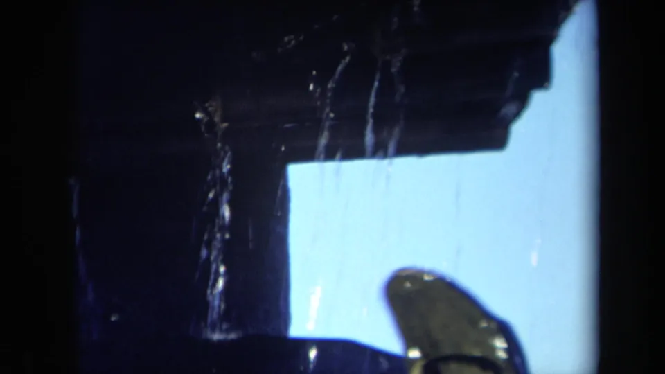 Close 8mm frame of water dripping from a dark mill edge, with bright blue sky or reflected light behind it.