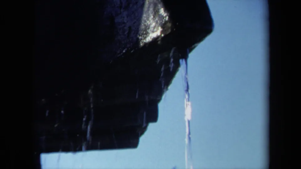 Close 8mm frame of a wet dark propeller blade, with water falling in a narrow stream against a clear blue sky.