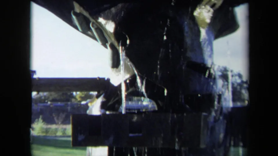 A close 1969 Sacramento home-movie view of water spilling over a dark tiered fountain against grass and sky.