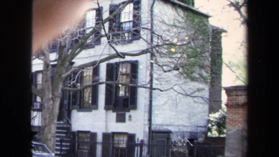 1970 Annapolis home-movie frame showing a white brick townhouse, black shutters, bare tree branches, and a dark foreground window frame.