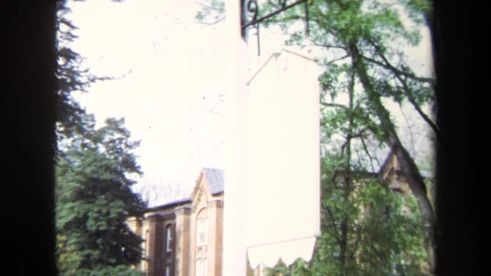 1970 Annapolis exterior view with a brick building, green trees, pale wall, and dark vertical foreground obstruction.