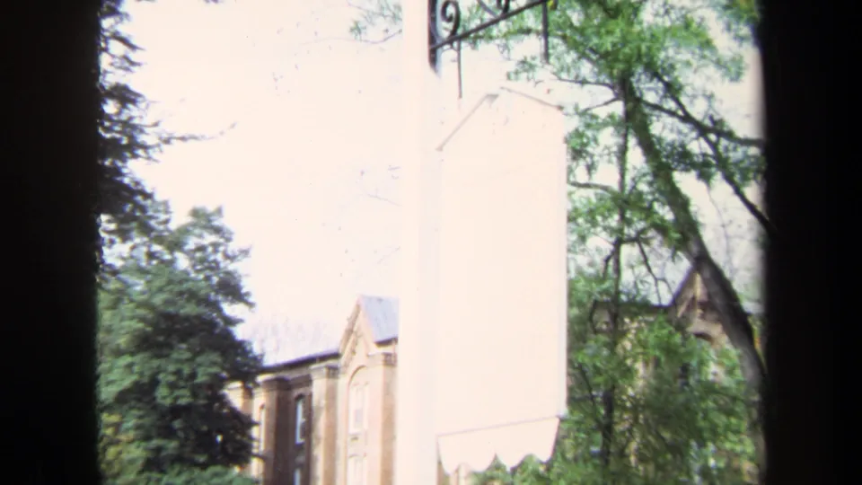 1970 Annapolis frame showing a brick building through trees beside a pale wall with a small white hanging sign.