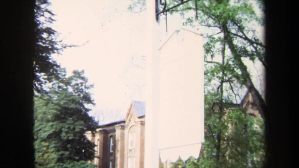1970 Annapolis home-movie view of a brick building and leafy trees, partially blocked by a pale wall and dark window frame.