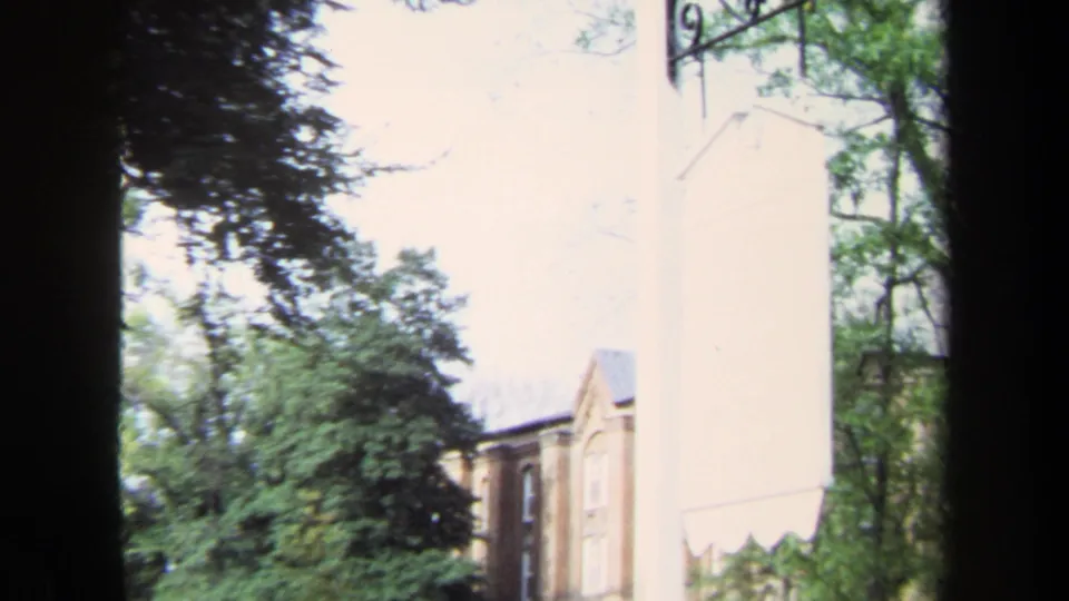 1970 Annapolis 8mm frame looking through trees toward a pale building wall and part of a brick house on landscaped grounds.