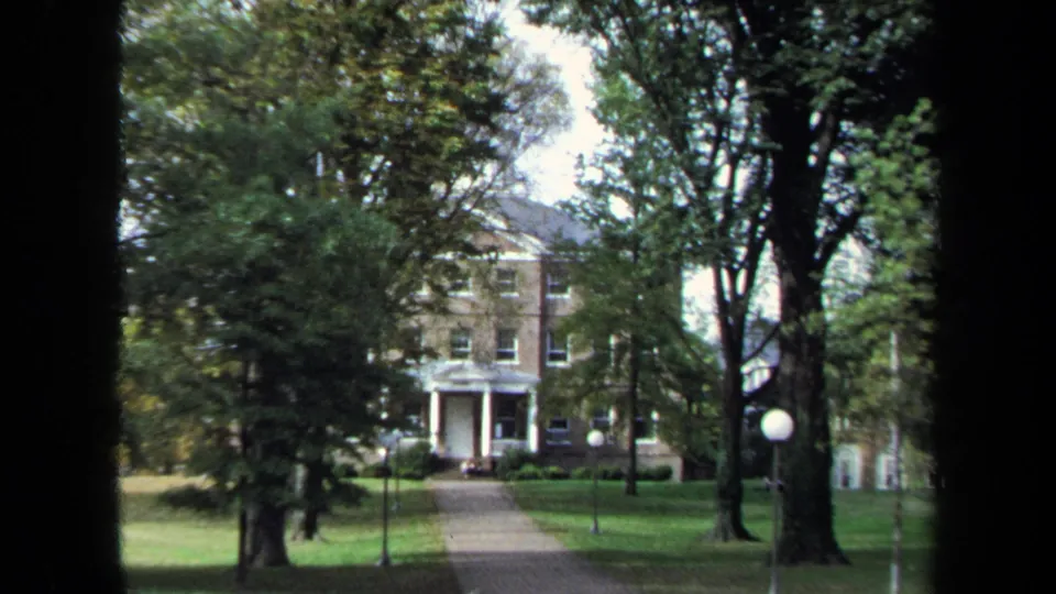 1970 Annapolis 8mm frame of a large brick house with a white-columned entrance, straight walkway, trees, lawn, and globe lamps.