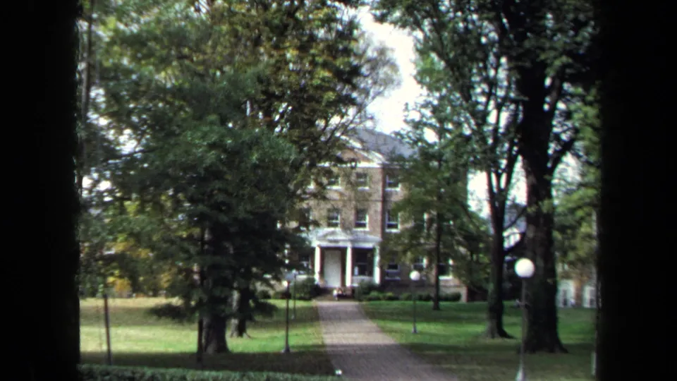 1970 Annapolis 8mm frame centered on a brick house facade with white entrance columns, a path, lawn, trees, and round lamp posts.