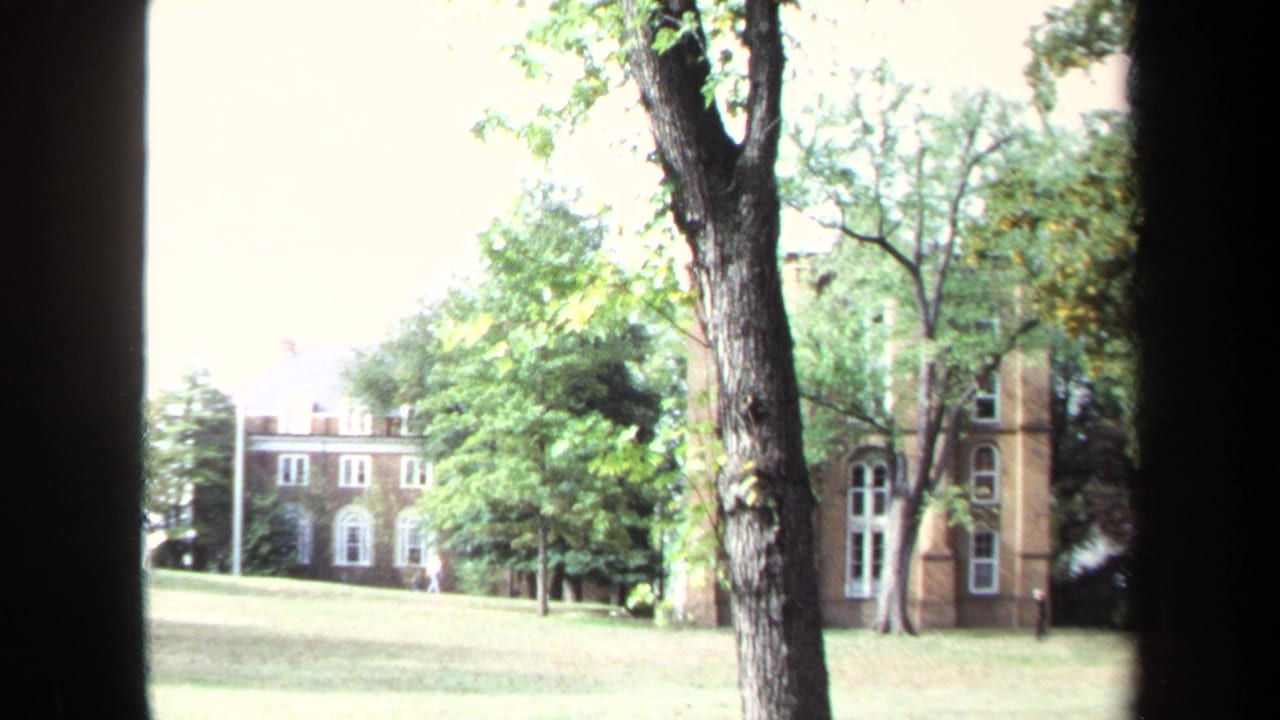 1970 Annapolis campus view with brick buildings, white-trimmed windows, grass, trees, and dark vertical foreground framing.